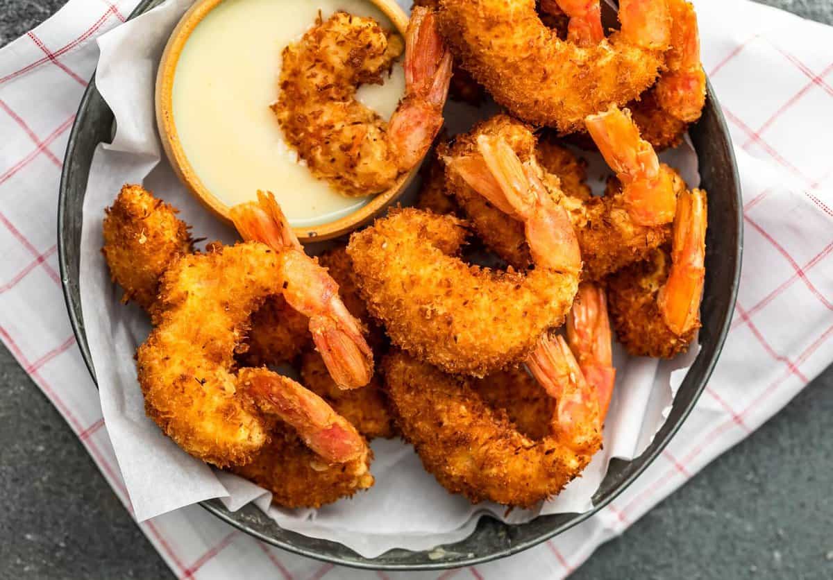 Coconut Shrimp is a crispy, crave-worthy appetizer that everyone will love. It's especially delicious when paired with the perfect coconut shrimp sauce: a Spicy Pina Colada Dipping Sauce! The two go hand in hand, a match made in heaven. And this coconut shrimp recipe is just as good as a main dish as it is as a party appetizer! a plate of coconut shrimp with a small bowl of pina colada dipping sauce.