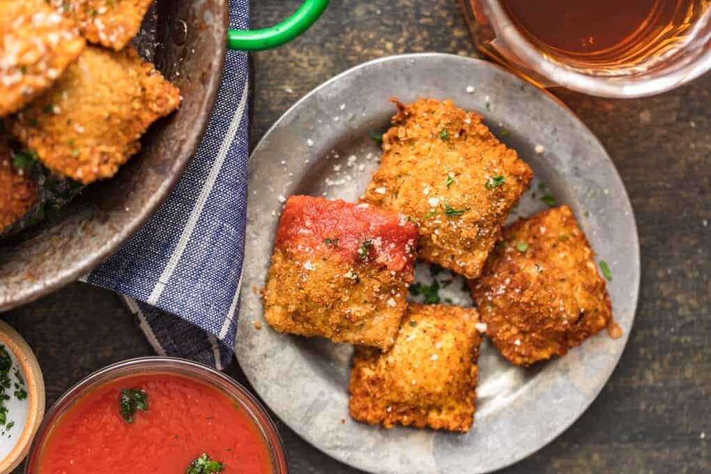 4 toasted ravioli on a gray plate, one is dipped in tomato sauce.