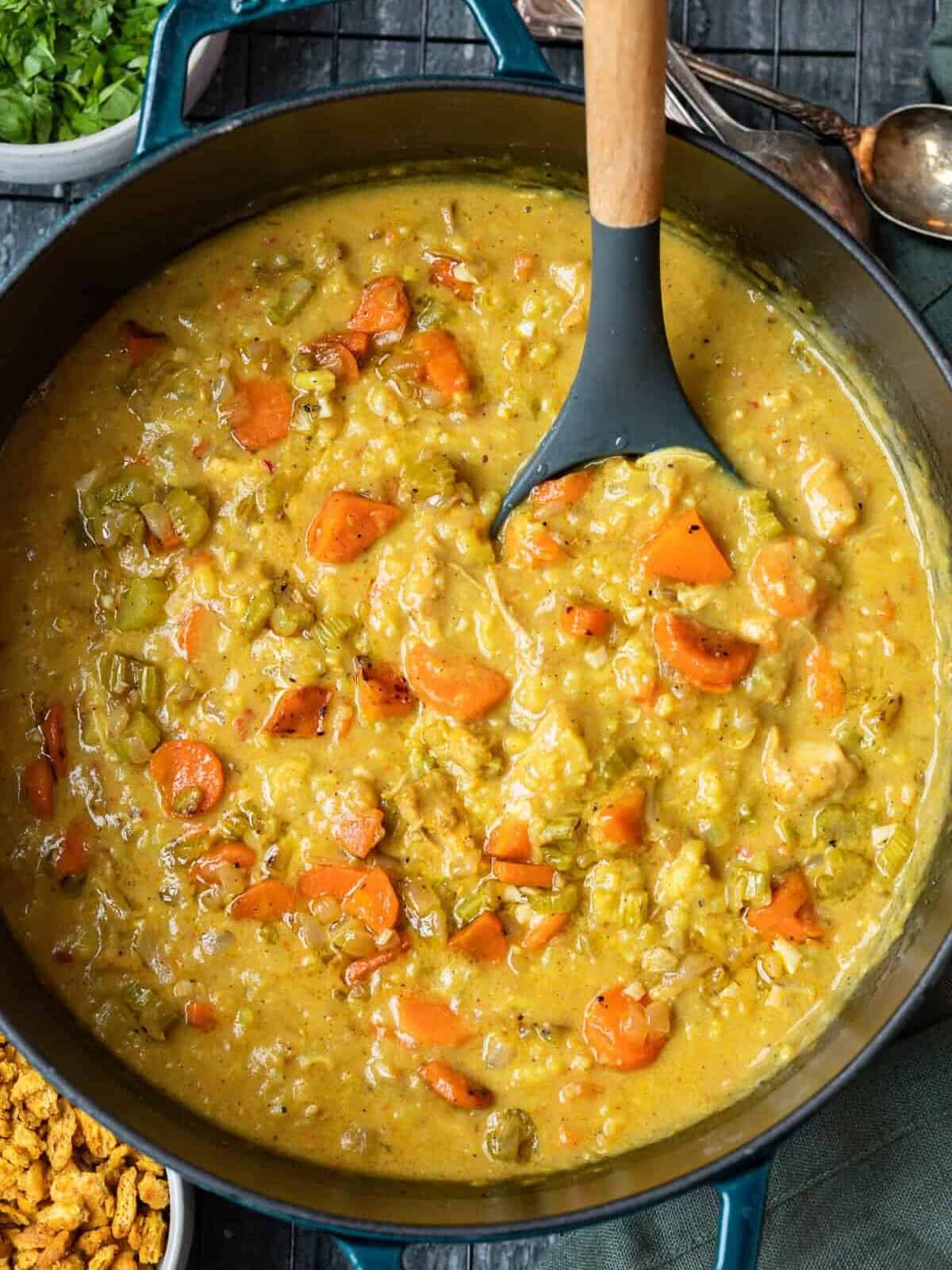 overhead view of Indian mulligatawny soup in a dutch oven with a serving spoon. Sliced carrots, celery, chicken, and rice are visible in the creamy curry base.