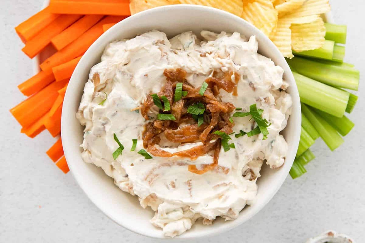 overhead image of french onion dip in a white bowl on a serving platter surrounded by chips, celery sticks, and carrot sticks