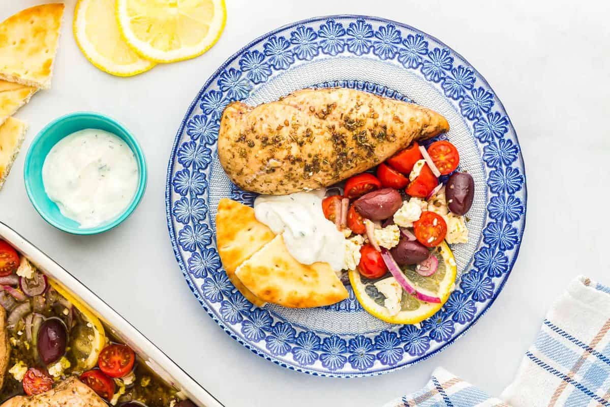overhead view of a baked greek chicken breast on a blue patterned plate with pita bread and yogurt.