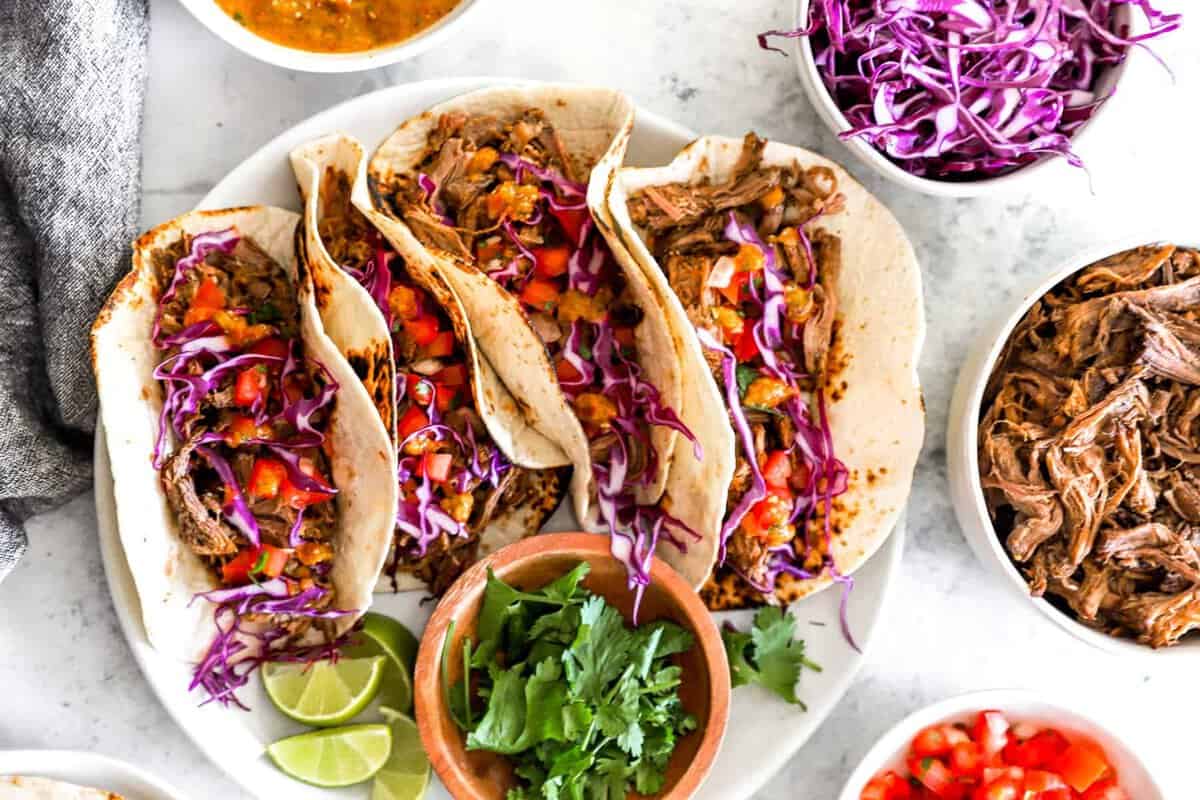 overhead view of 4 barbacoa tacos on a white plate with bowls of toppings around them.