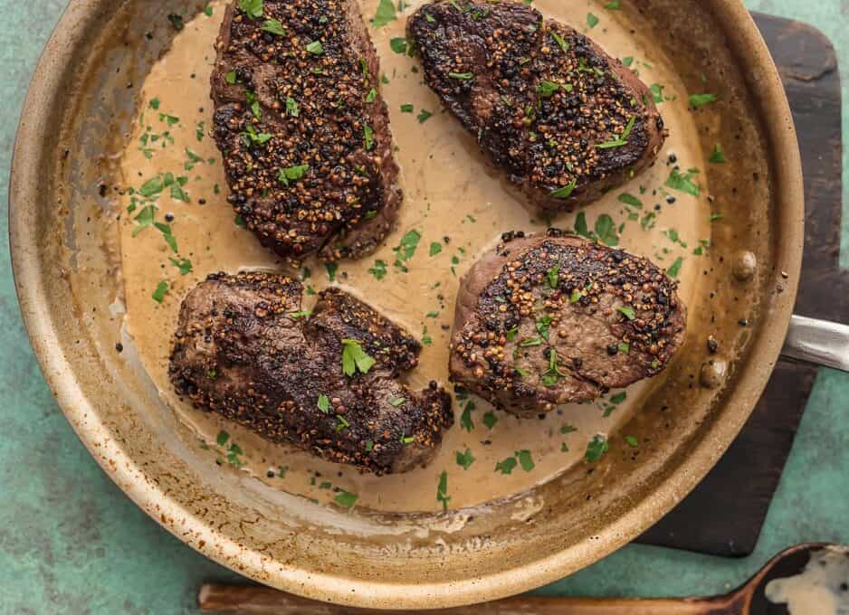 four peppercorn steaks sitting in a skillet with brandy cream sauce.