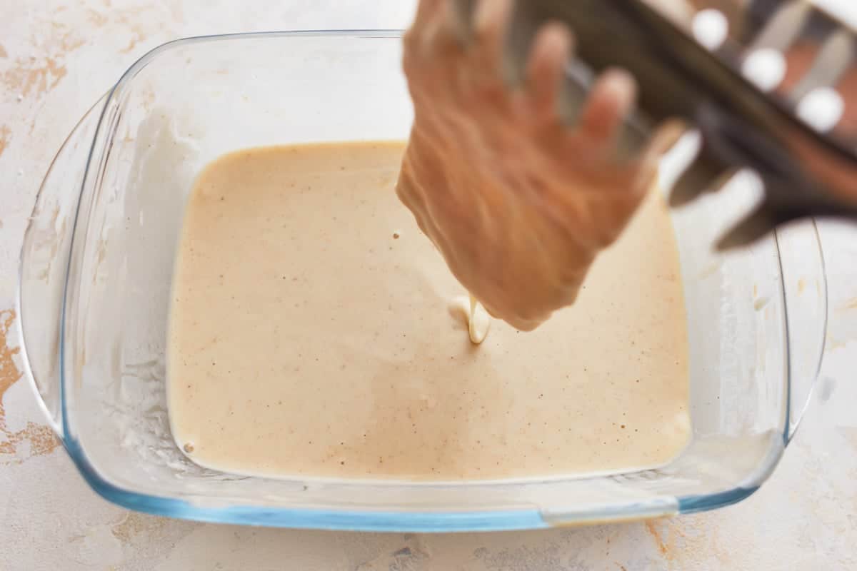 Tongs dipping a fish fillet in beer batter in a glass dish.