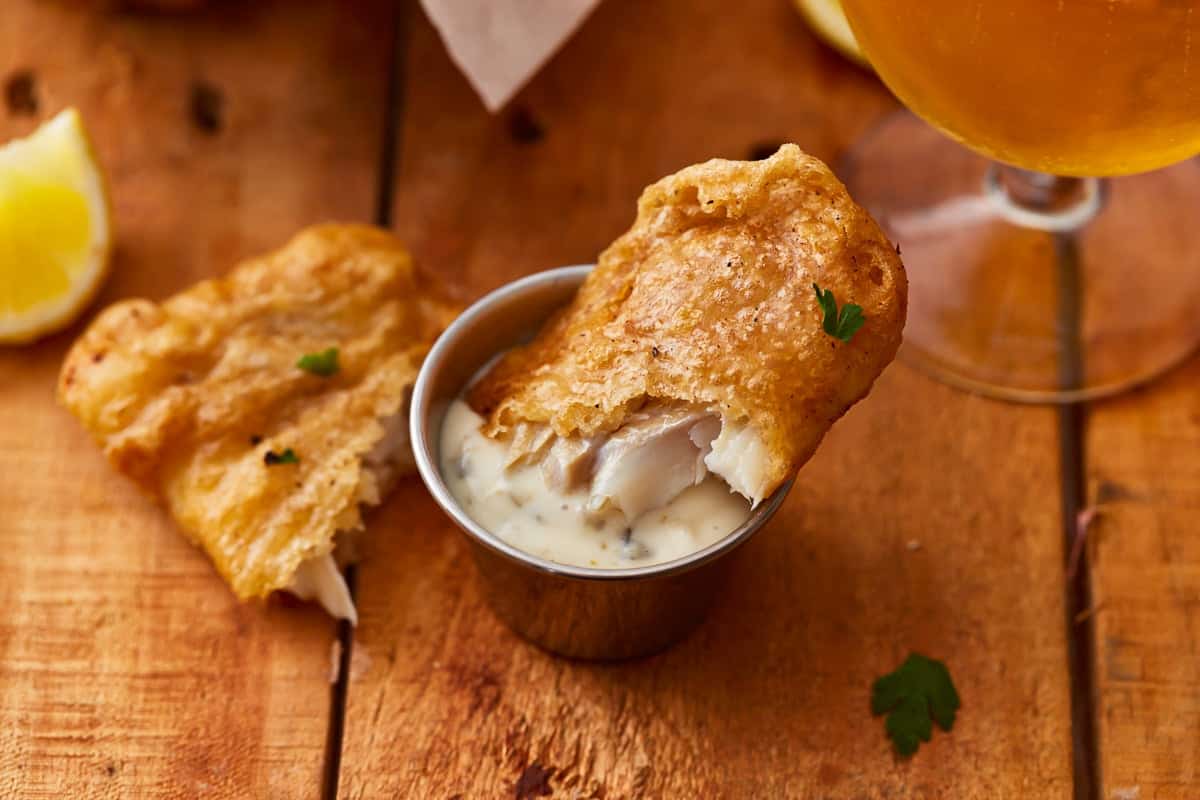 Beer battered fish dipped in tartar sauce on a wooden table next to a glass of beer.