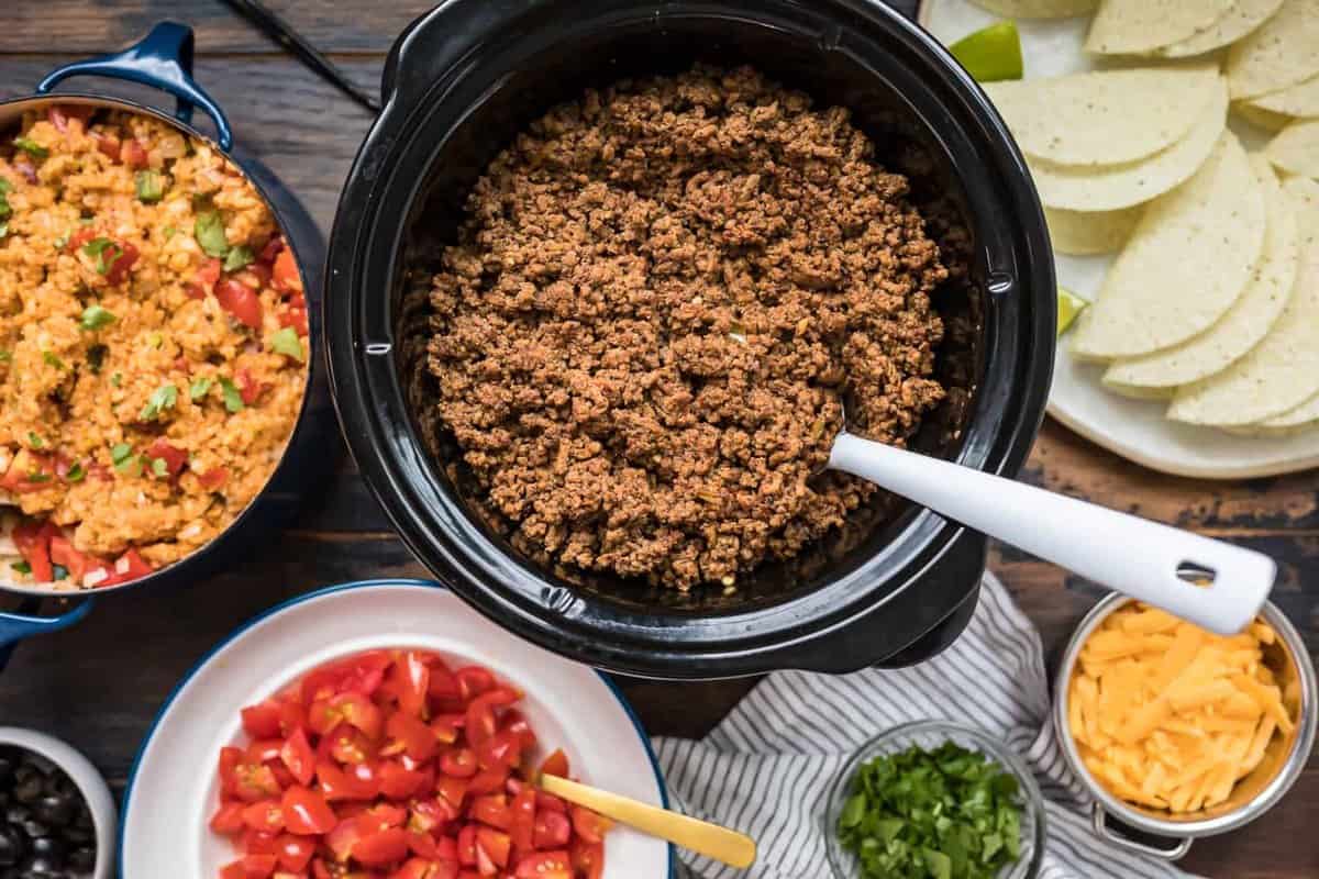 crockpot taco meat in a slow cooker with a spoon and Mexican rice, a plate of taco shells, a bowl of pico de gallo, a bowl of cheese and bowl of cilantro.