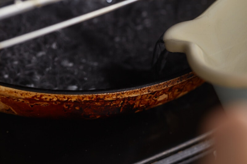 A person pouring milk into a pan while a French baguette sits nearby.