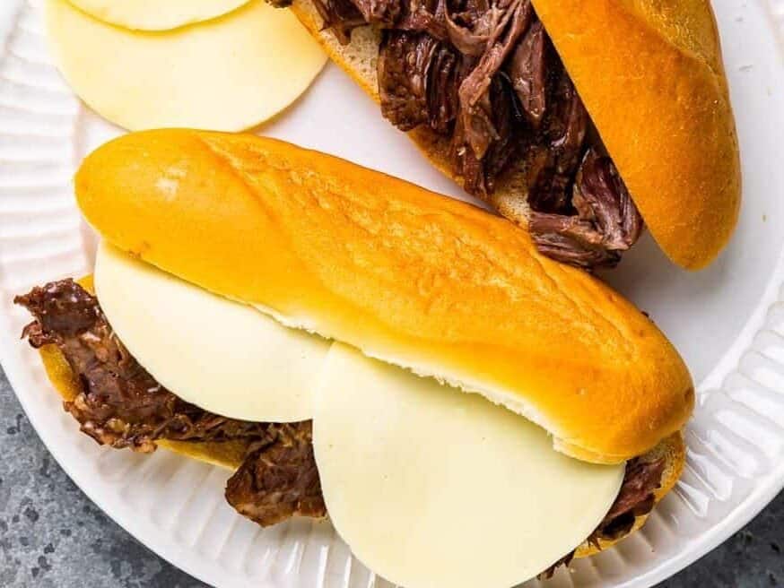an assembled french dip sandwich on a white plate next to a french dip sandwich without any cheese.