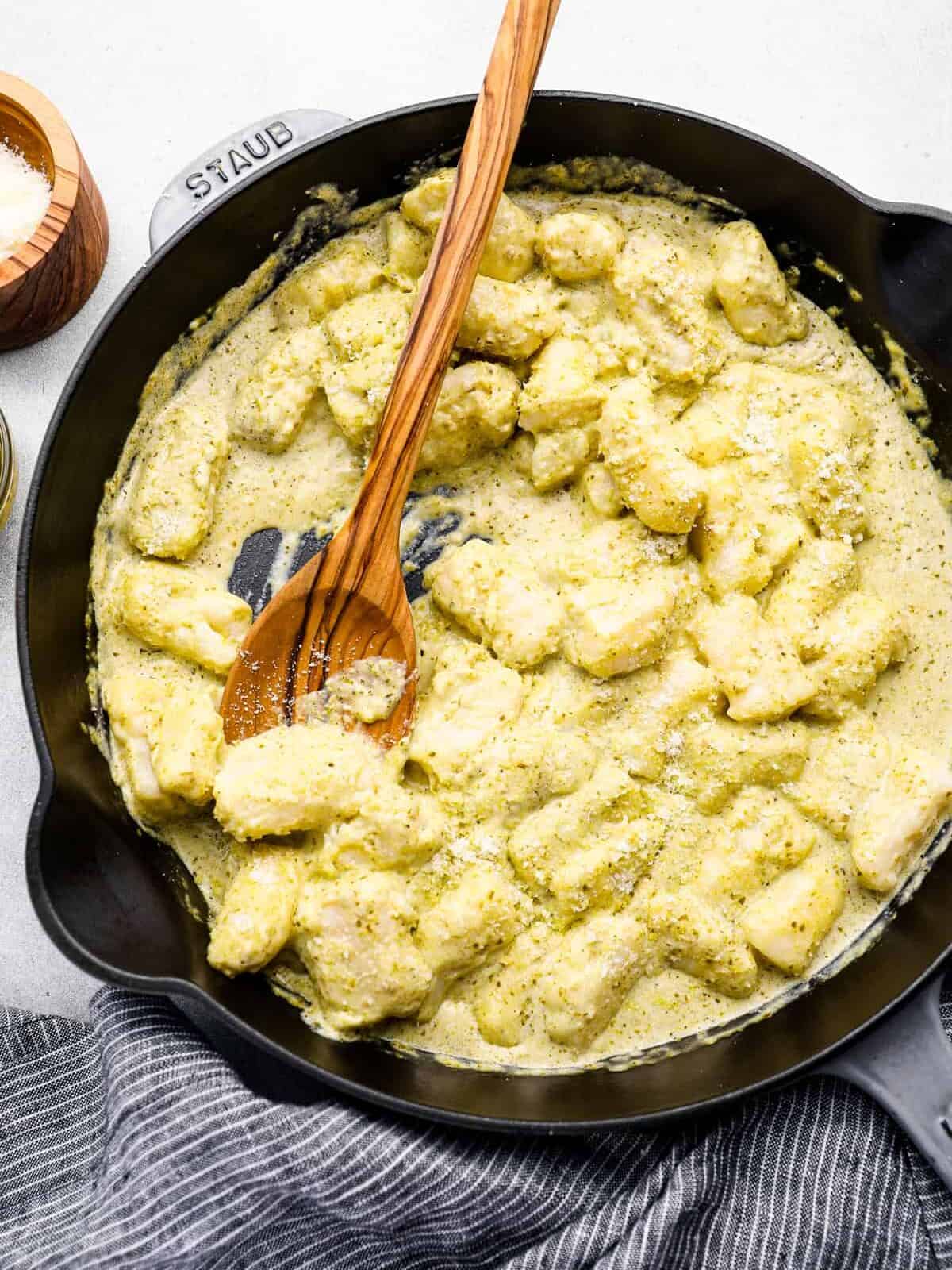 overhead view of homemade gnocchi in pesto cream sauce in a cast iron skillet with a wooden spoon.