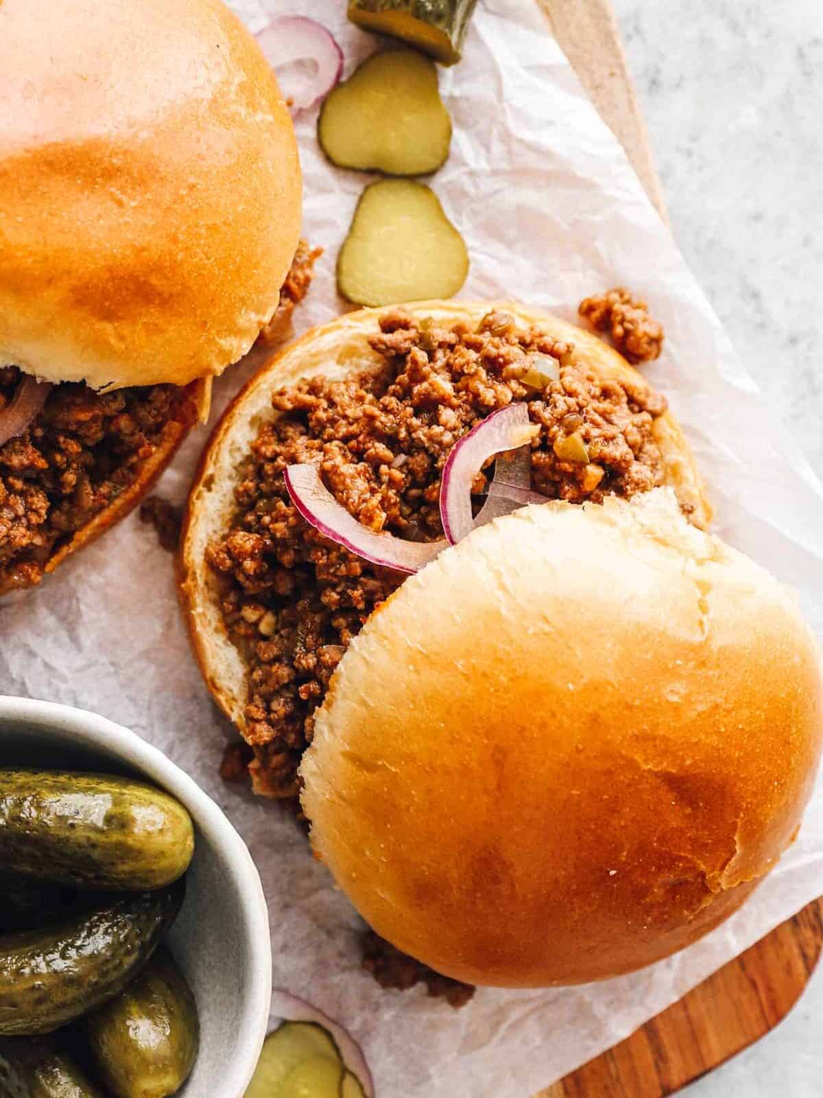 close up view of 2 sloppy joe sandwiches on a wooden cutting board with pickles.