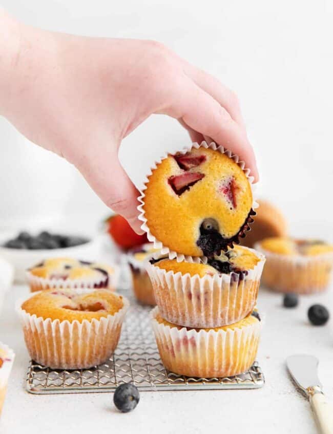 a hand holding a wildberry muffin in front of a wire rack full of wildberry muffins.