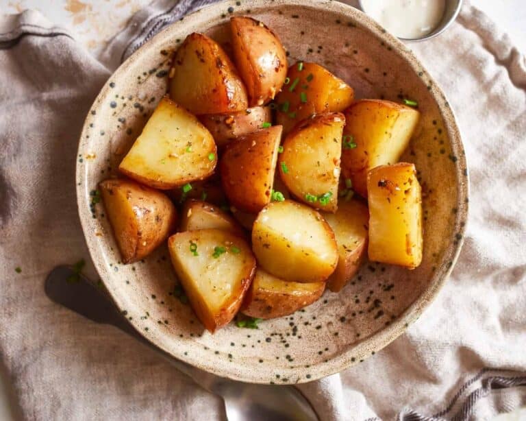 A ceramic bowl filled with golden-brown roasted baby potatoes garnished with chopped chives. The bowl sits on a beige cloth napkin, and a fork is partially visible to the side.