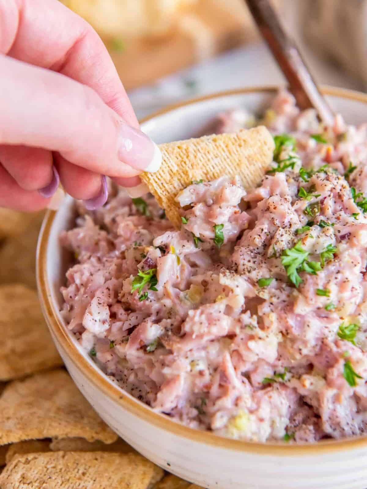 a hand dipping a triscuit into a bowl of ham salad.