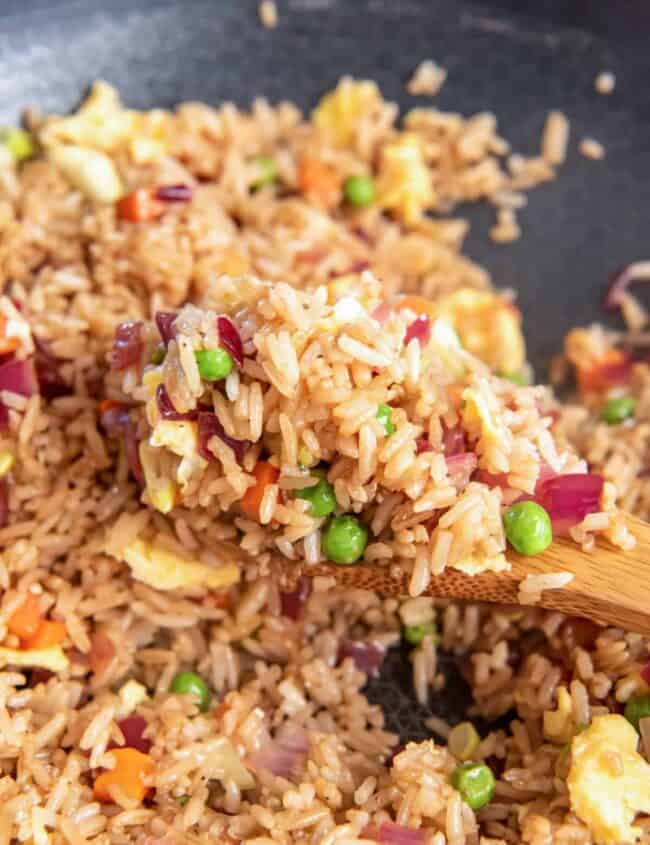 close up view of a wooden spoon lifting a scoop of vegetable fried rice from a wok.