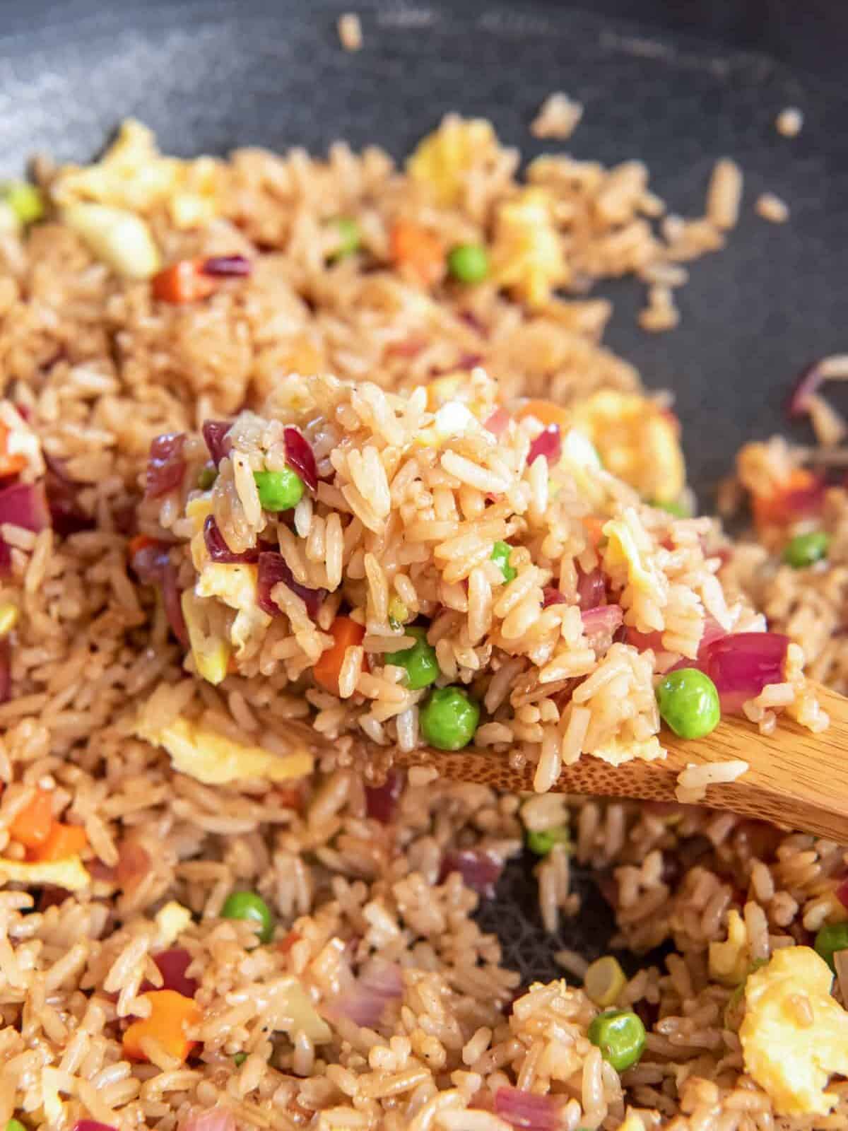 close up view of a wooden spoon lifting a scoop of vegetable fried rice from a wok.