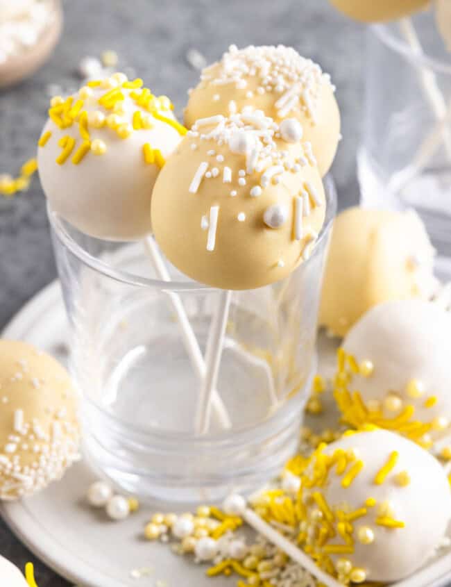 lemon cake pops arranged standing up in a glass, with more cake bites scattered around a plate, with sprinkles and edible pearls as decoration.