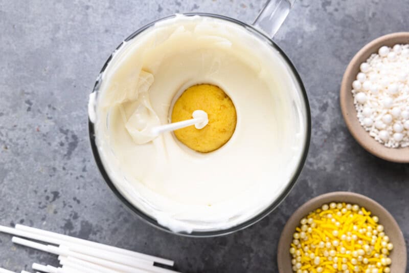 a lemon cake pop on a stick, dipped into a bowl of melted white chocolate.