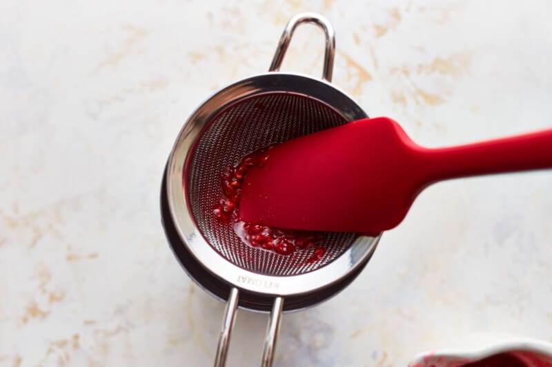 A rubber spatula pressing muddled raspberries through a mesh sieve.