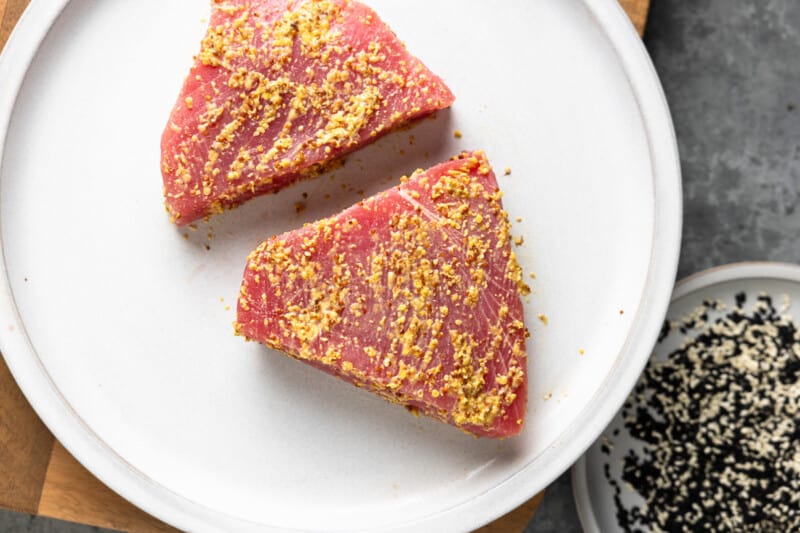 two raw tuna steaks coated in Dijon mustard, set on a white plate.
