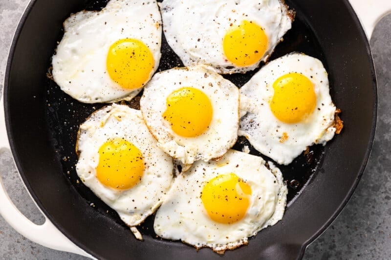 six fried eggs cooking in a skillet.