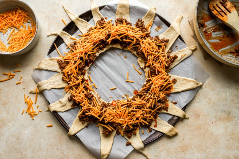 cheese and ground beef laid over crescent roll triangles arranged around a bowl on a baking sheet.