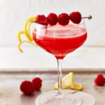 A red-hued raspberry lemon drop cocktail set on a coaster, with lemon slices and raspberries scattered around it.