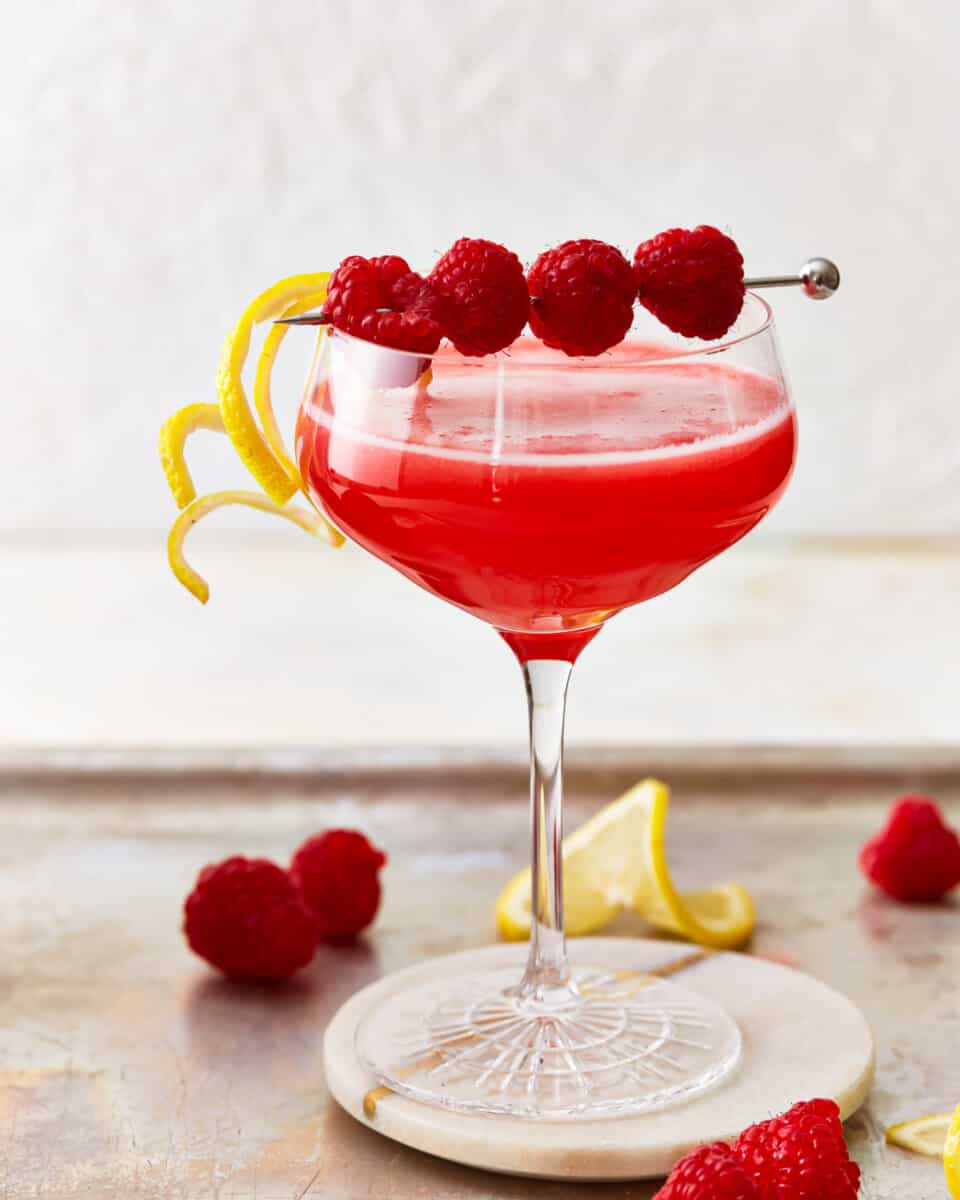 A red-hued raspberry lemon drop cocktail set on a coaster, with lemon slices and raspberries scattered around it.