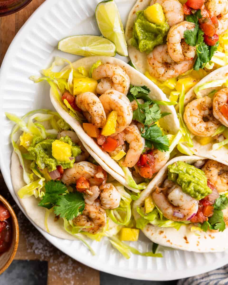 Shrimp and pineapple slaw tacos on a white plate, garnished with lime wedges and cilantro.