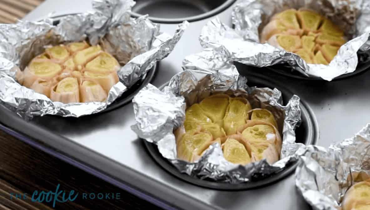 Roasted garlic bulbs in aluminum foil in a muffin tray.