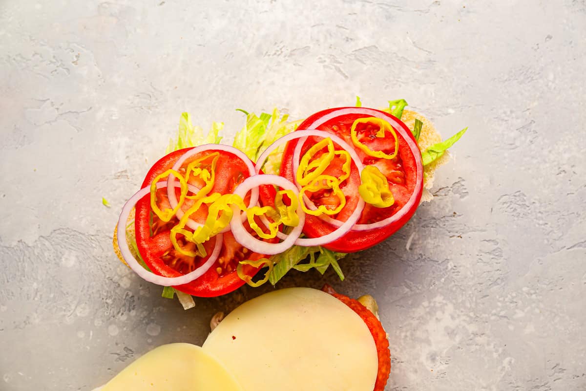 meats, cheeses, lettuce, tomato, red onion, and pepperoncini layered on the bottom half of a sub roll.
