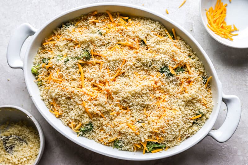 unbaked chicken divan in a white casserole dish, topped with shredded cheese, broccoli, and breadcrumbs.
