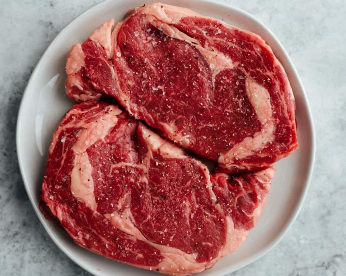 Two raw seasoned rib eye steaks on a white plate. - 9