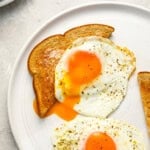 2 sunny side up eggs on a white plate, one draped over a slice of toast.