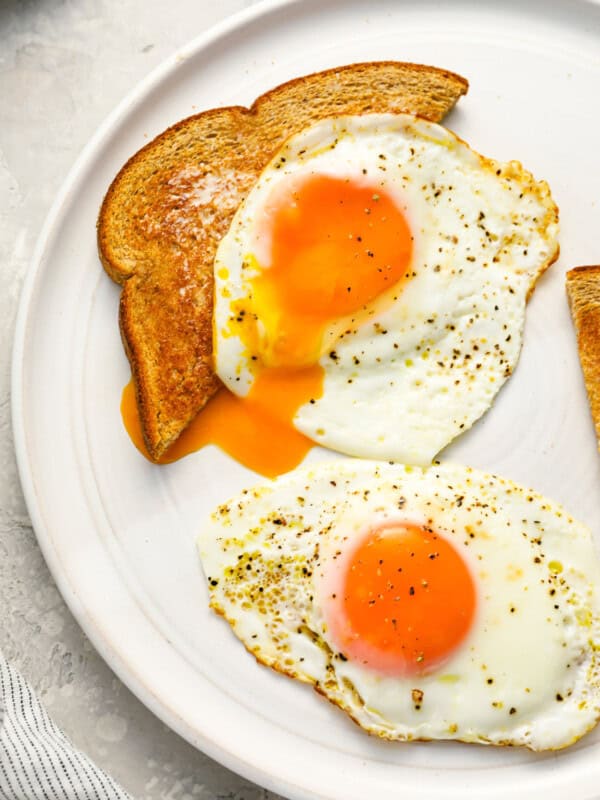2 sunny side up eggs on a white plate, one draped over a slice of toast.
