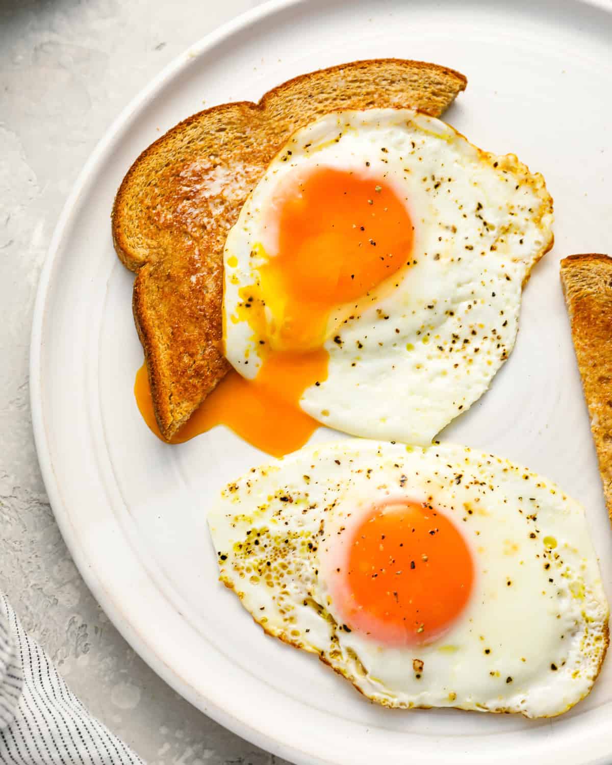 2 sunny side up eggs on a white plate, one draped over a slice of toast.