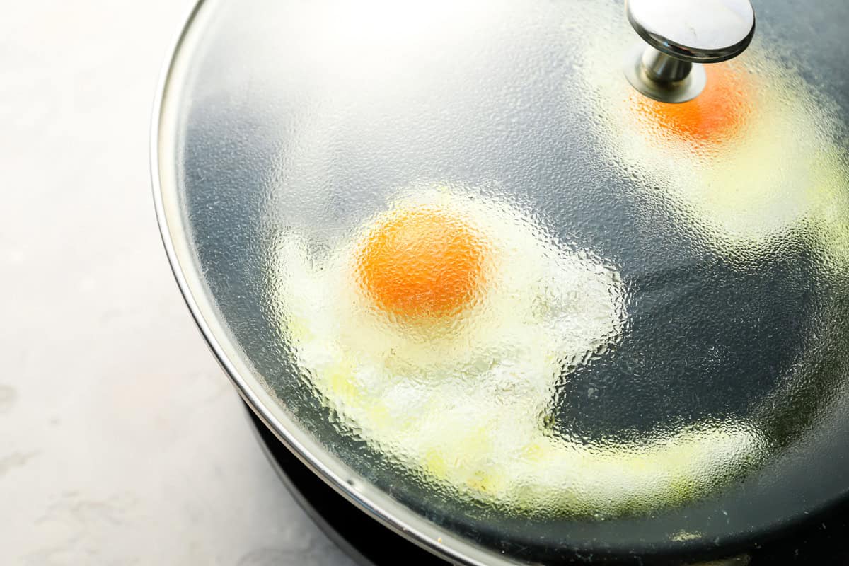 steaming sunny side up eggs in a nonstick pan covered with a glass lid.