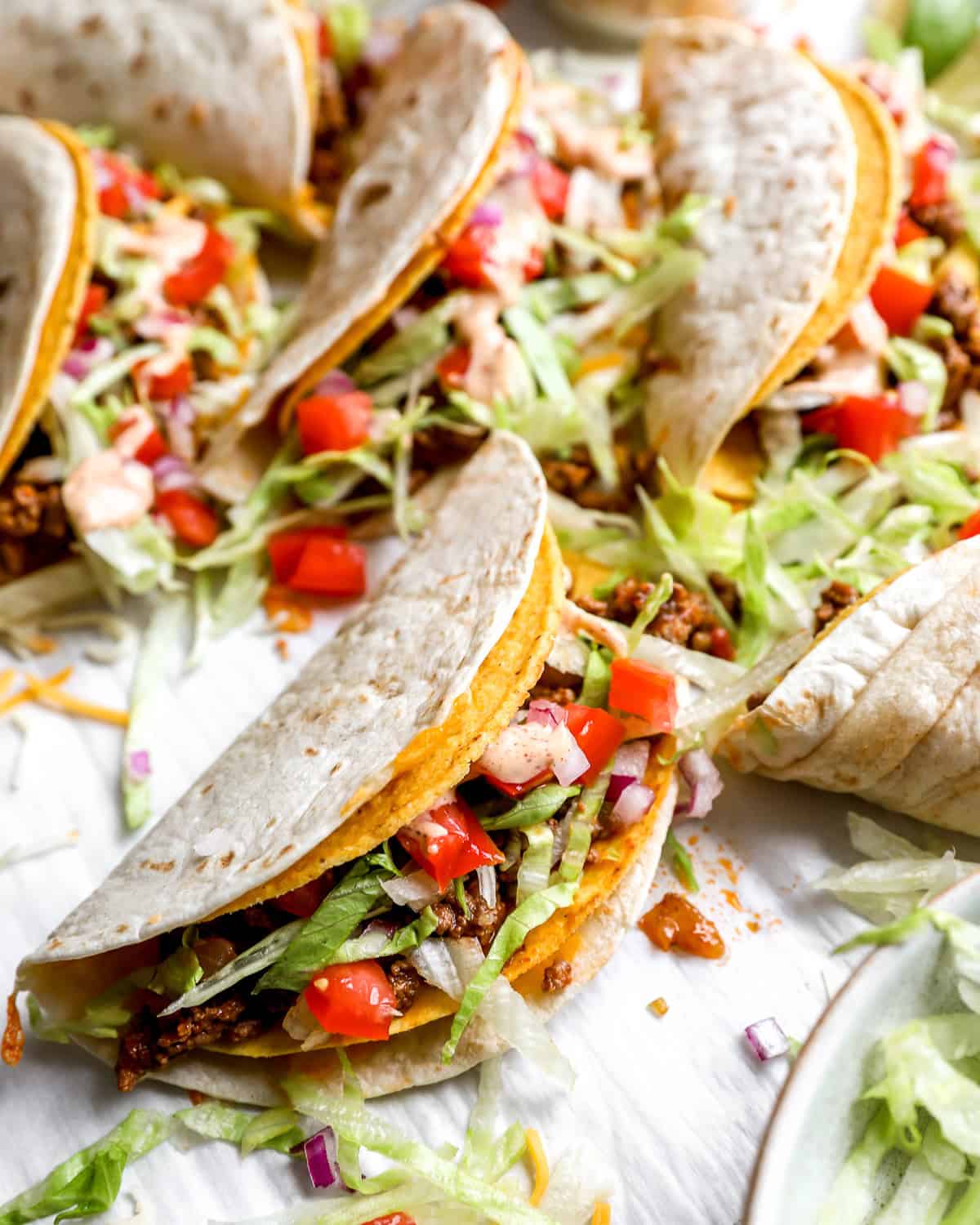 Several tacos filled with ground beef, lettuce, tomatoes, and cheese, placed in a row on a white surface