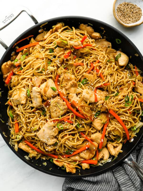 A pan filled with stir-fried noodles, chicken pieces, red bell peppers, and green onions, topped with sesame seeds. A small bowl of sesame seeds is placed beside the pan.