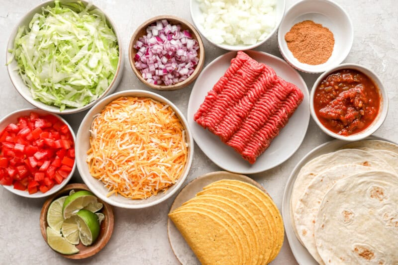 ingredient picture with shredded cheese, ground beef. red and yellow onions, shredded lettuce, limes, tomatoes, hard taco shells, flour tortillas, taco seasoning