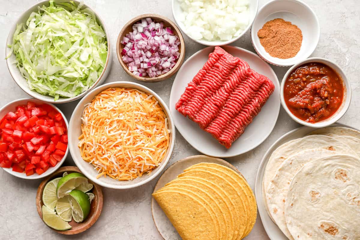 ingredient picture with shredded cheese, ground beef. red and yellow onions, shredded lettuce, limes, tomatoes, hard taco shells, flour tortillas, taco seasoning