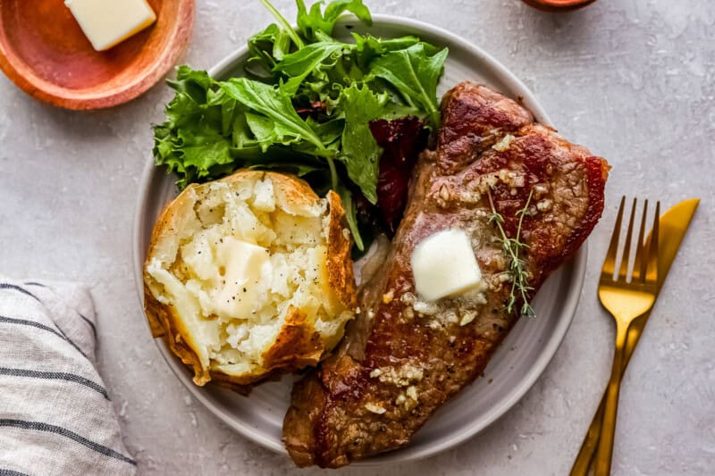 new york strip steak topped with butter on a plate wirh a baked potato and mixed greens