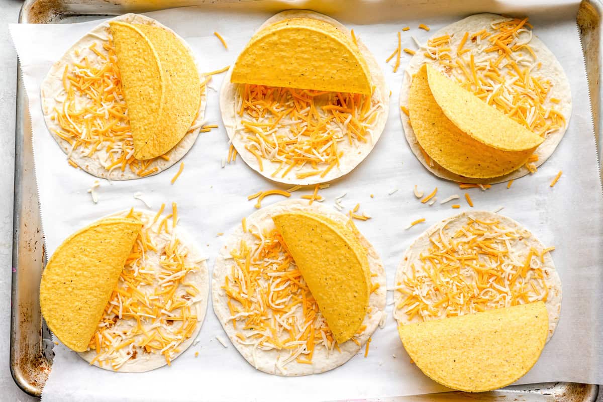 flour tortillas topped with shredded cheese and hard taco shells on a lined baking sheet