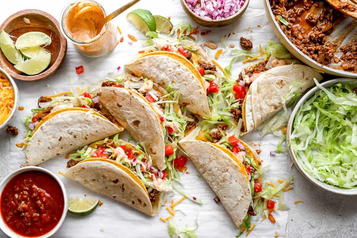 Several tacos filled with ground beef, lettuce, tomatoes, and cheese, placed in a row on a white surface with ingredients surrounding them
