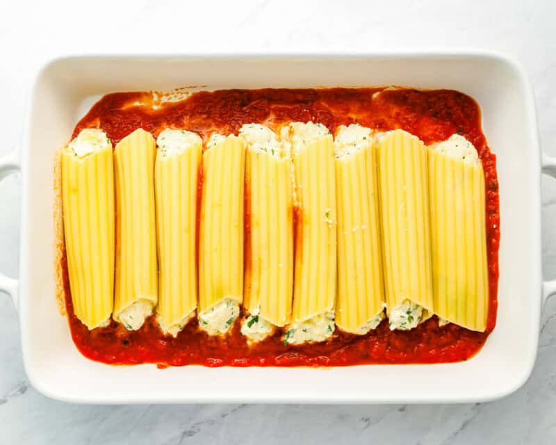 filled manicotti lined up on top of marinara sauce in white baking dish
