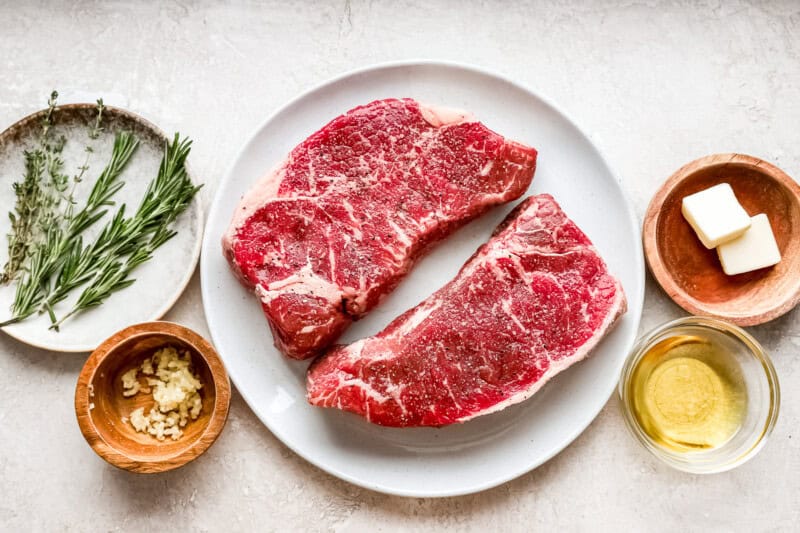 ingredients laid out: two raw new york strip steaks, fresh thyme and rosemary sprigs, minced garlic cloves, avocado oil, butter