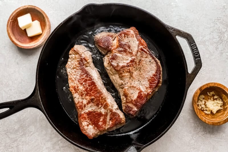 two new york steak strips being browned in a cast iron pan