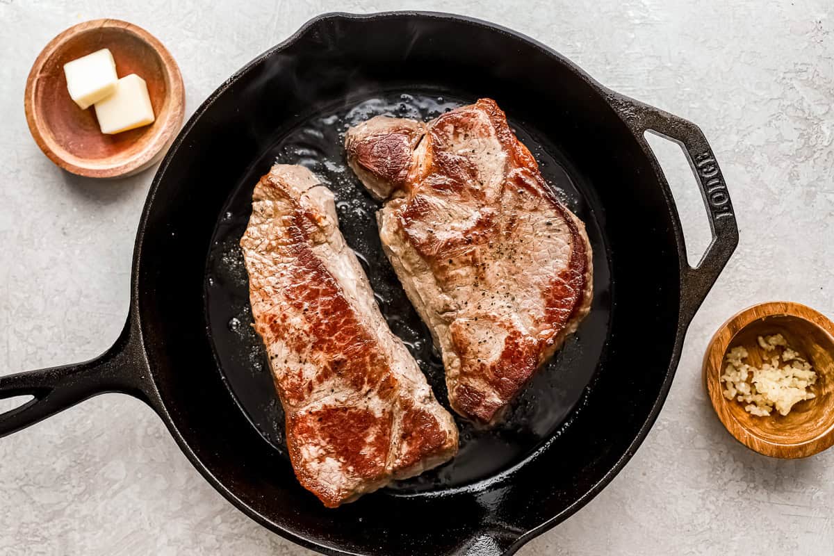 two new york steak strips being browned in a cast iron pan