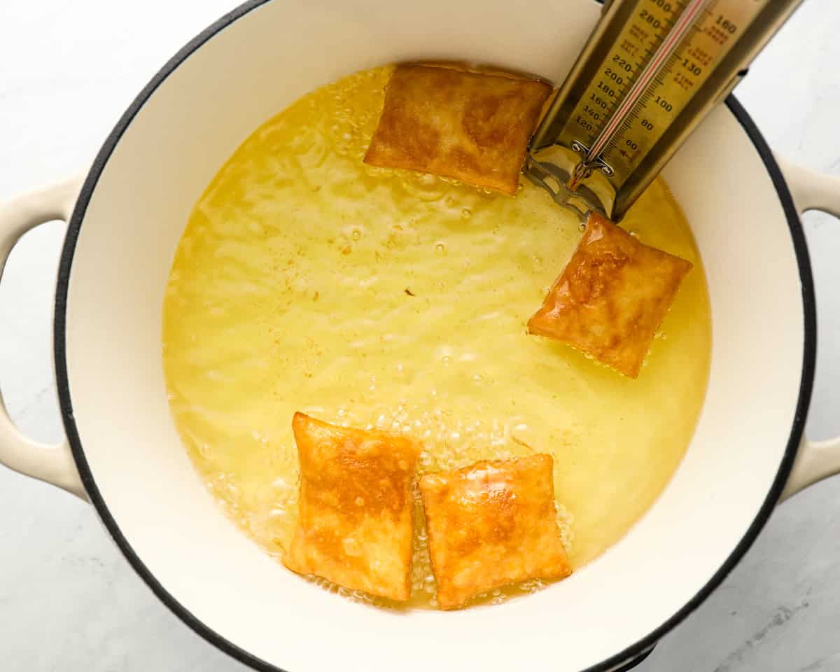Sopapillas frying in a pot of oil, turning golden brown, with a thermometer clipped to the side to monitor the temperature.