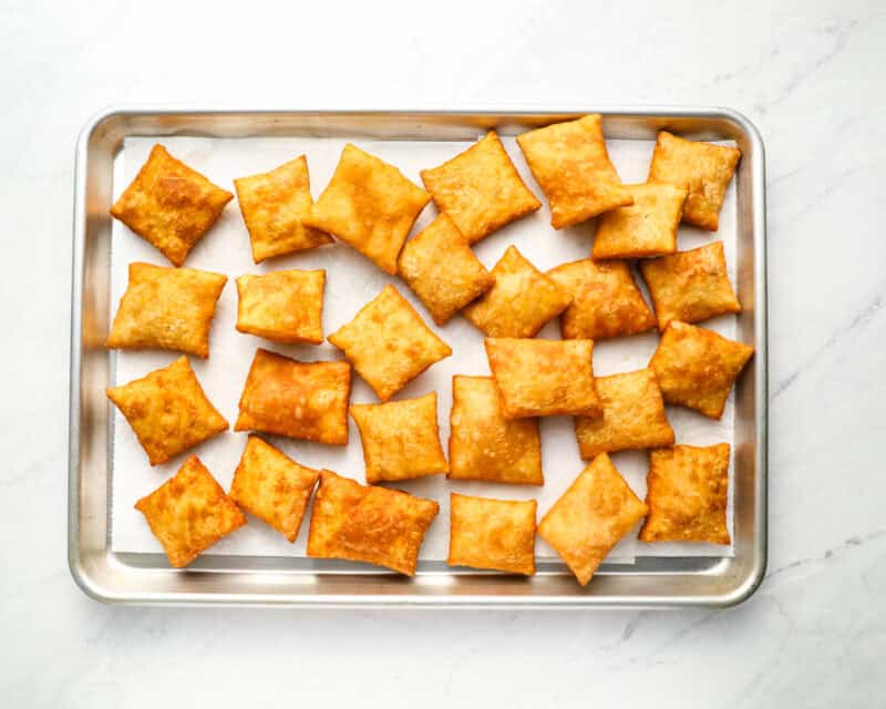 A baking tray lined with paper towels holding freshly fried sopapillas, golden and crispy, cooling after frying.