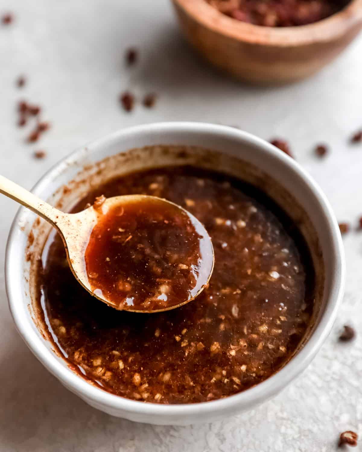 A close-up view of a small bowl filled with homemade Szechuan sauce, featuring visible minced garlic and chili flakes, with a golden spoon resting in the sauce.